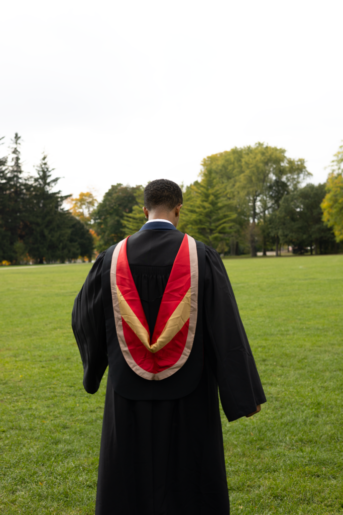 Person in a graduation gown with a red and gold stole standing on grass with trees in the background. The photo is from Guelph Grad Photos. The background is the University of Guelph Campus.