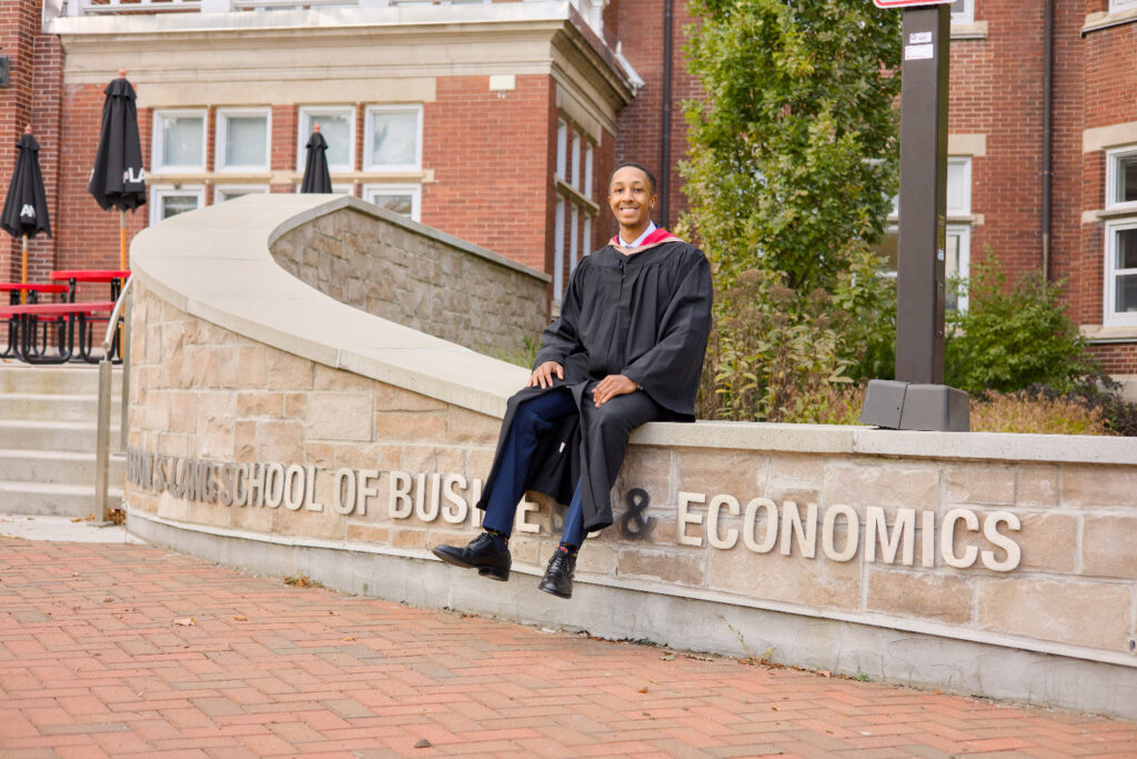 A professional photo of a Guelph grad on the University of Guelph campus in a cap and gown, sitting on a ledge with 'Economics' and 'School of Business' signs in the background. near the University of Guelph's LANG Building. The photo is from Guelph Grad Photos.