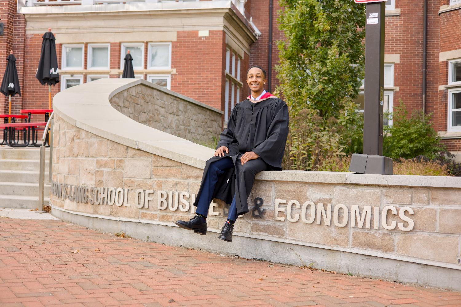 A professional photo of a Guelph grad on the University of Guelph campus in a cap and gown, sitting on a ledge with 'Economics' and 'School of Business' signs in the background. near the University of Guelph's LANG Building. The photo is from Guelph Grad Photos.