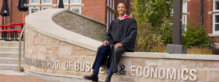 a Guelph grad in graduation gown sitting on a stone wall with 'LANG School of Business & Economics' inscription.