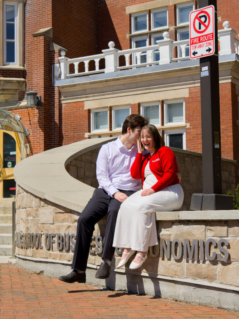 A professional photo of two Guelph grads on the University of Guelph campus, sitting on a ledge in front of a building with 'School of Business Economics' signage. The photo is from Guelph Grad Photos. The background is the LANG Building on the University of Guelph Campus.