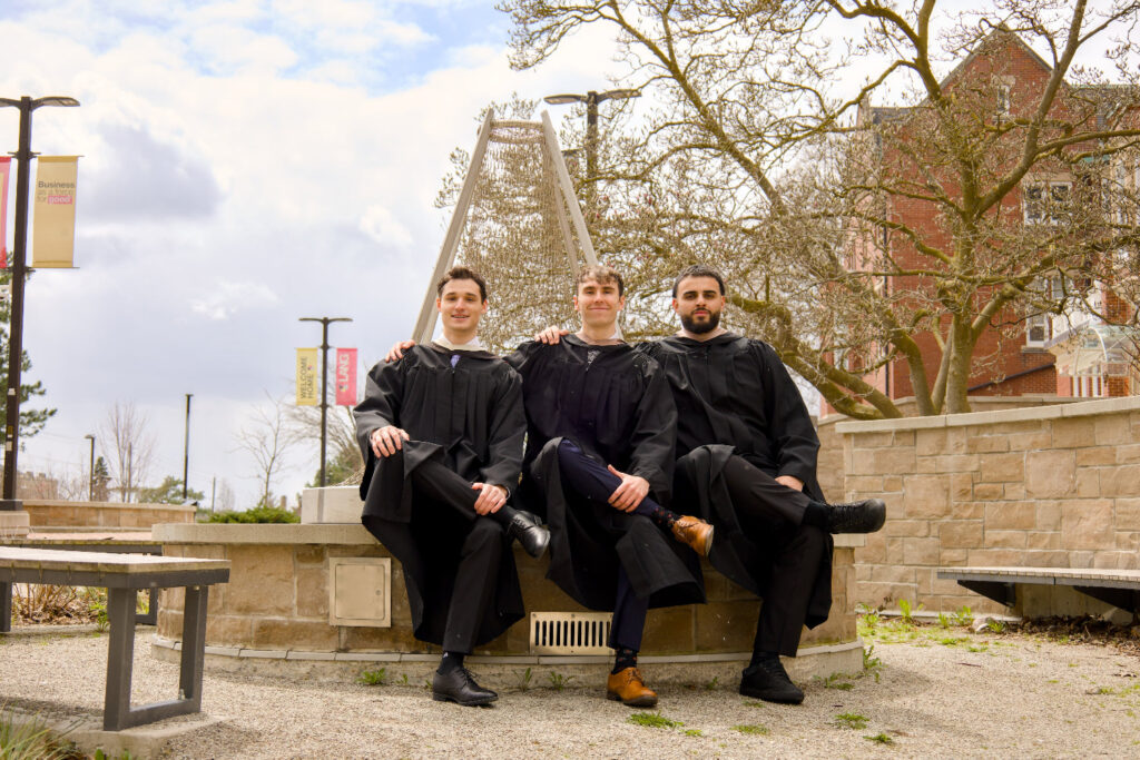 Three individuals in graduation gowns sitting on a bench outdoors with a building and trees in the background. The photo is from Guelph Grad Photos. The background is the University of Guelph Campus.