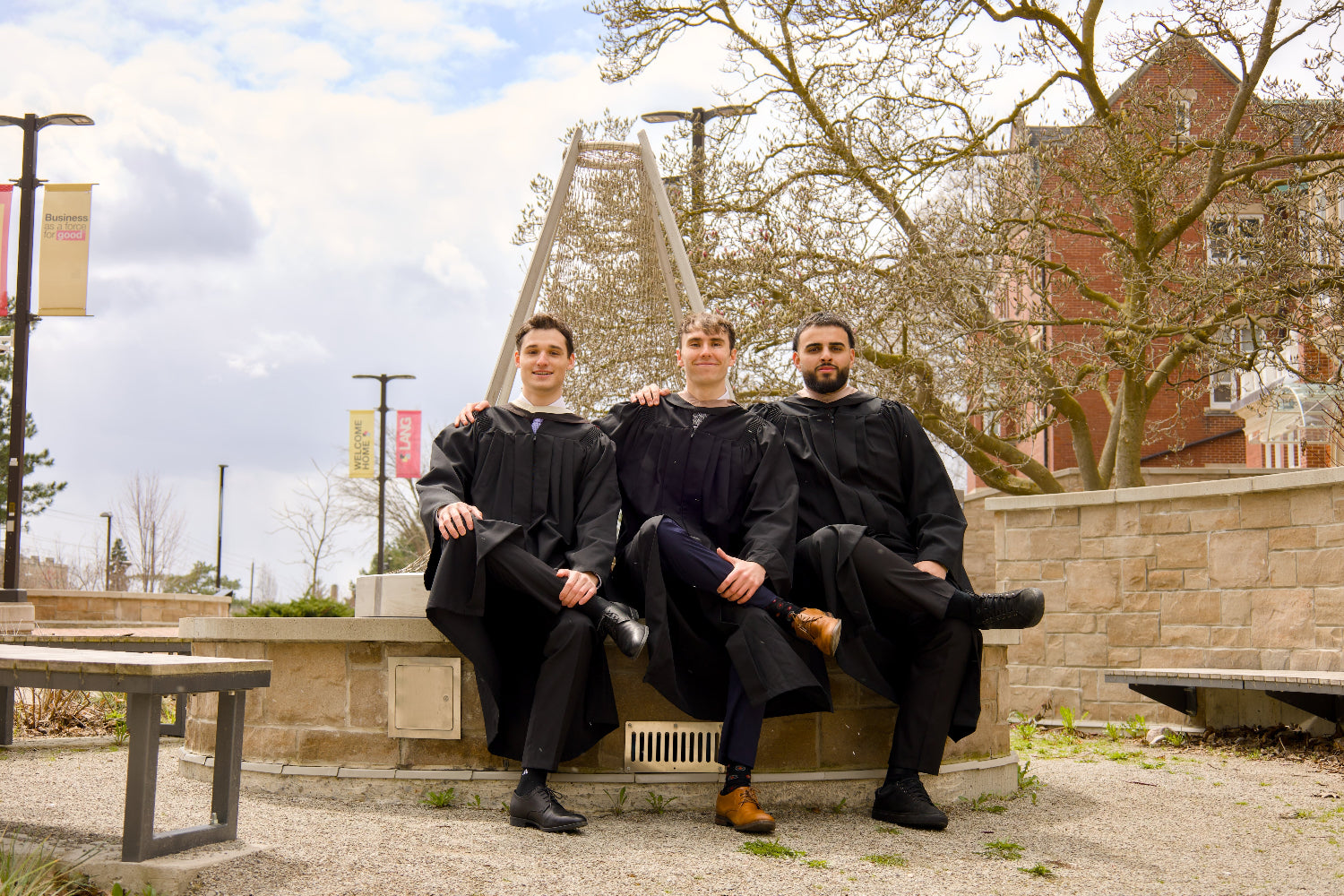 Three individuals in graduation gowns sitting on a bench outdoors with a building and trees in the background. The photo is from Guelph Grad Photos. The background is the University of Guelph Campus.