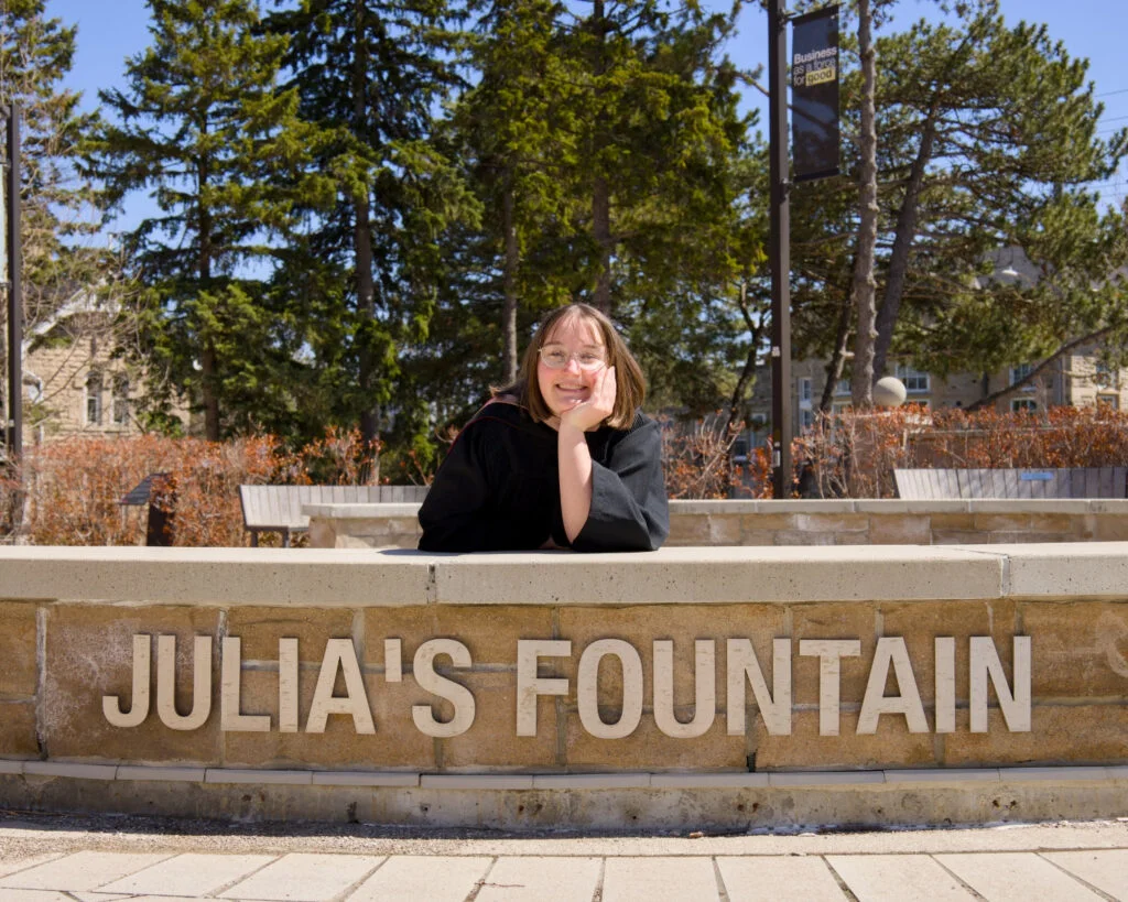 Person posing behind a fountain named 'Julia's Fountain' with trees and buildings in the background. The photo is from Guelph Grad Photos. The background is the University of Guelph Campus.