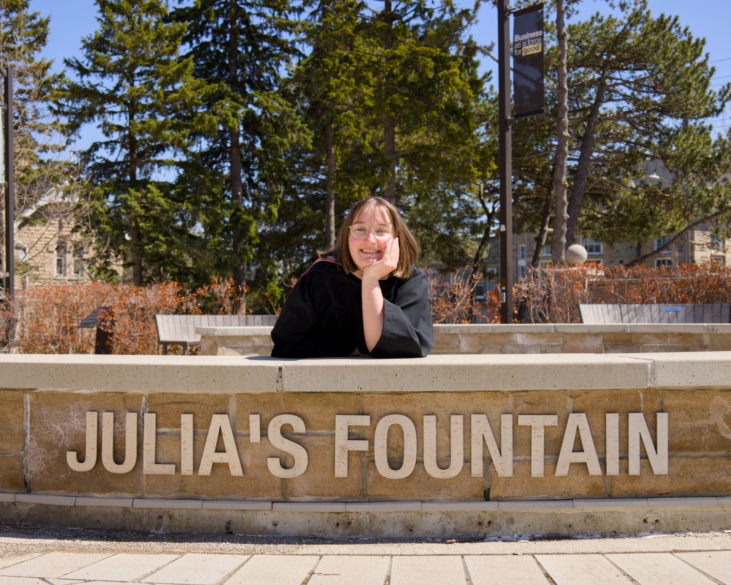 Person posing behind a fountain named 'Julia's Fountain' with trees and buildings in the background. The photo is from Guelph Grad Photos. The background is the University of Guelph Campus.