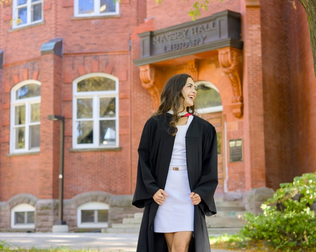Graduate in cap and gown standing in front of a brick building with 'Navye Hall Library' sign.