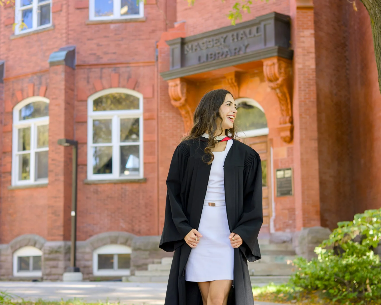 A University of Guelph graduate in cap and gown standing in front of the library building