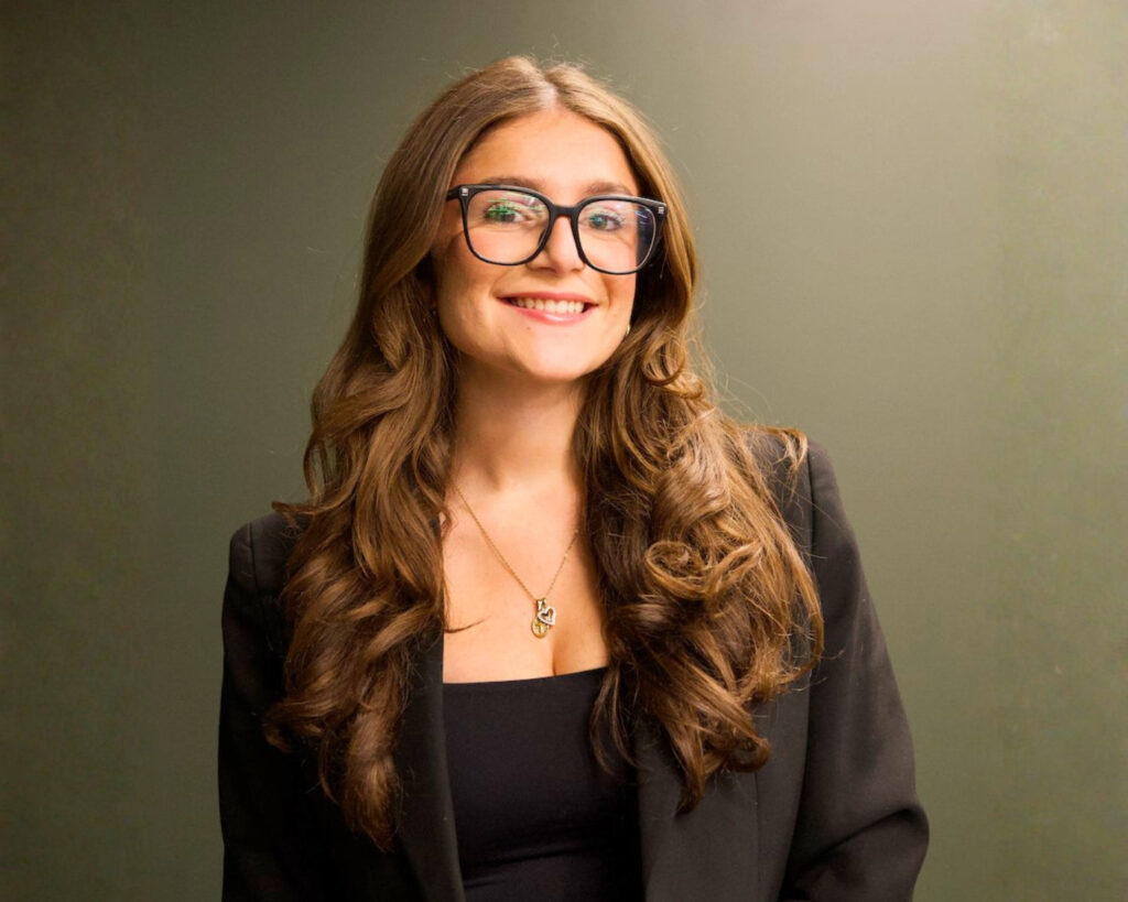 A Professional LinkedIn photo of a Guelph Grad, a Woman wearing glasses and a dark blazer against a green background