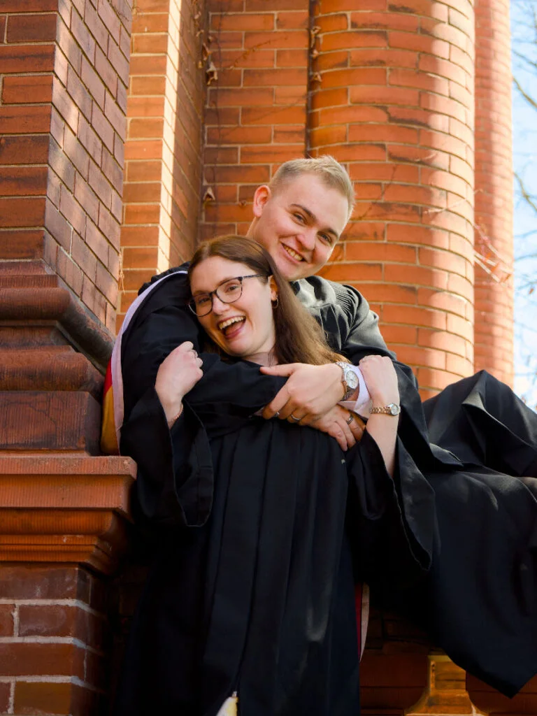 Two people in graduation gowns hugging in front of a brick building. The photo is from Guelph Grad Photos. The background is the University of Guelph Campus.