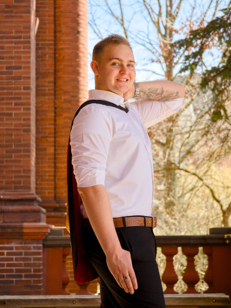 Man wearing a white shirt and black cape standing outdoors with a brick building and trees in the background The photo is from Guelph Grad Photos. The background is the University of Guelph Campus.