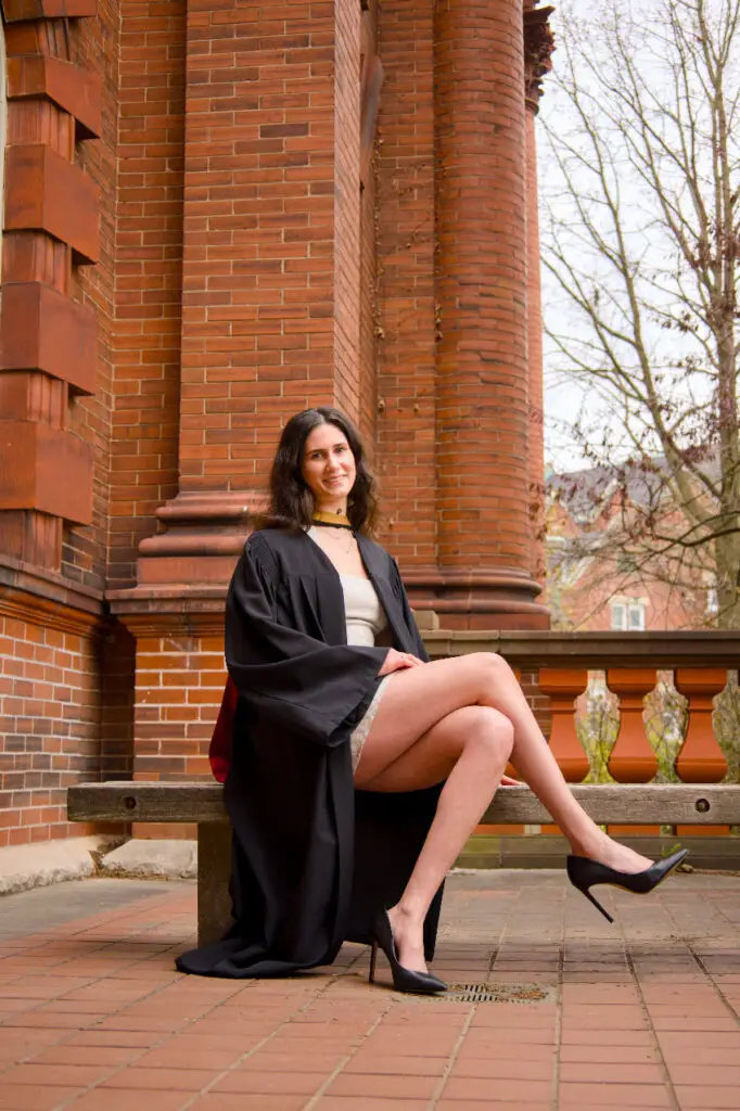 Woman in a black graduation gown sitting on a bench in front of a brick building. The photo is from Guelph Grad Photos. The background is the University of Guelph Campus.