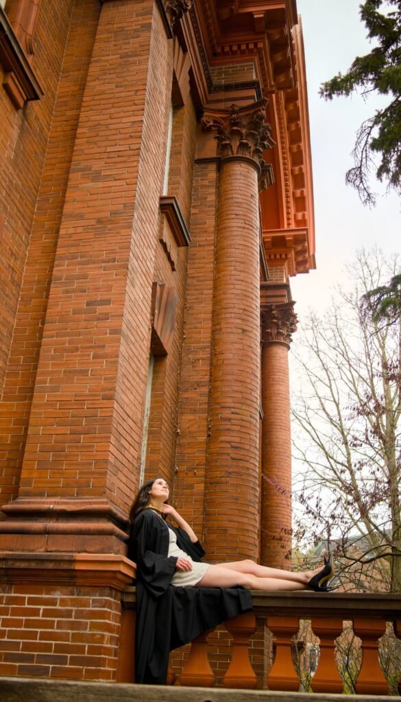 Woman in a black dress sitting on a stone ledge against a brick building with columns. The photo is from Guelph Grad Photos. The background is the University of Guelph Campus.