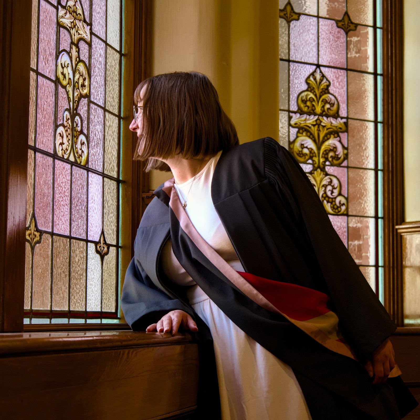 Person in academic gown standing by a stained glass window. The photo is from Guelph Grad Photos. The background is the University of Guelph Campus.