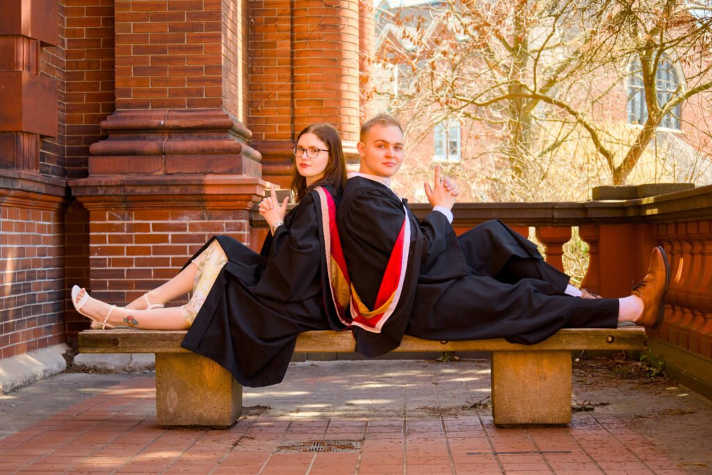 Two graduates in caps and gowns sitting on a bench outdoors. The photo is from Guelph Grad Photos. The background is the University of Guelph Campus.