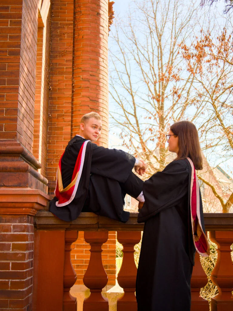 Two University of Guelph grads in graduation gowns sitting on a balcony with trees and a clear sky in the background.