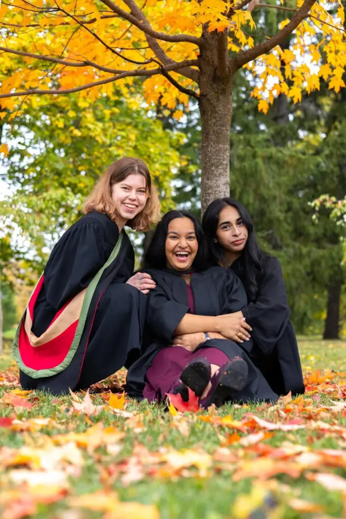 Three women in graduation gowns sitting on the ground with autumn leaves and trees in the background. The photo is from Guelph Grad Photos. The background is the University of Guelph Campus.