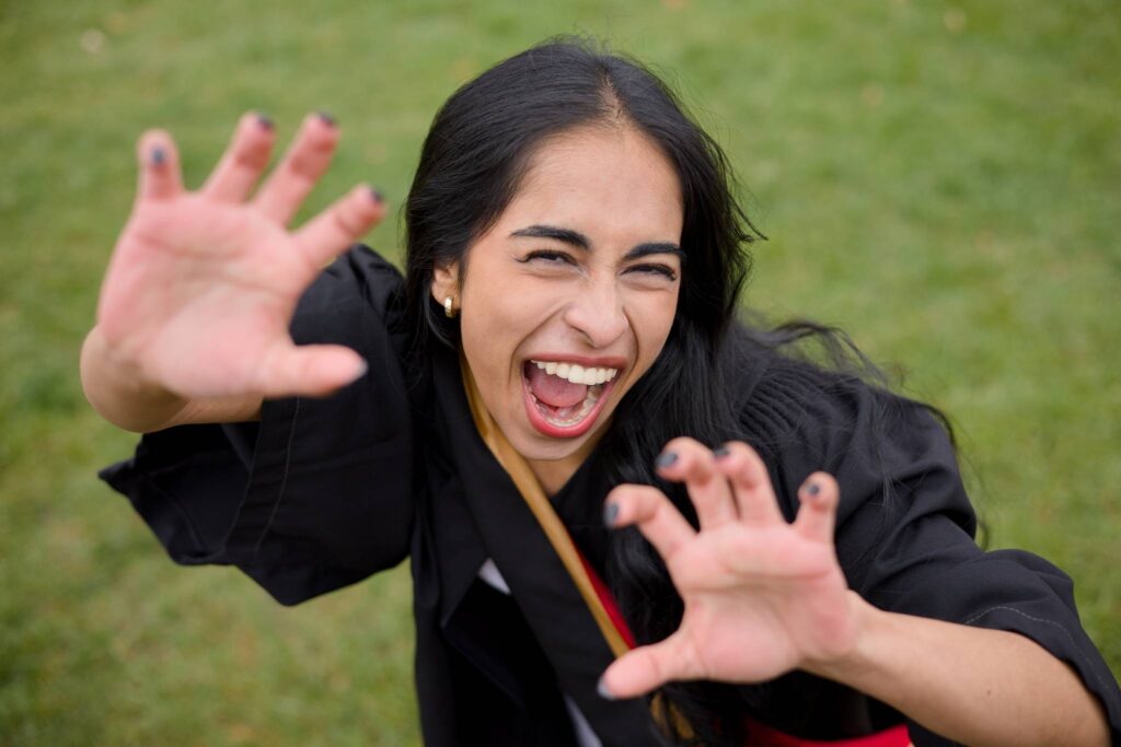 Woman in a black graduation gown with hands outstretched against a green background. The photo is from Guelph Grad Photos. The background is the University of Guelph Campus.