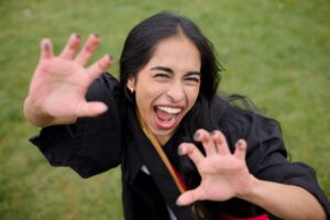 Woman in a black graduation gown with hands outstretched against a green background. The photo is from Guelph Grad Photos. The background is the University of Guelph Campus.
