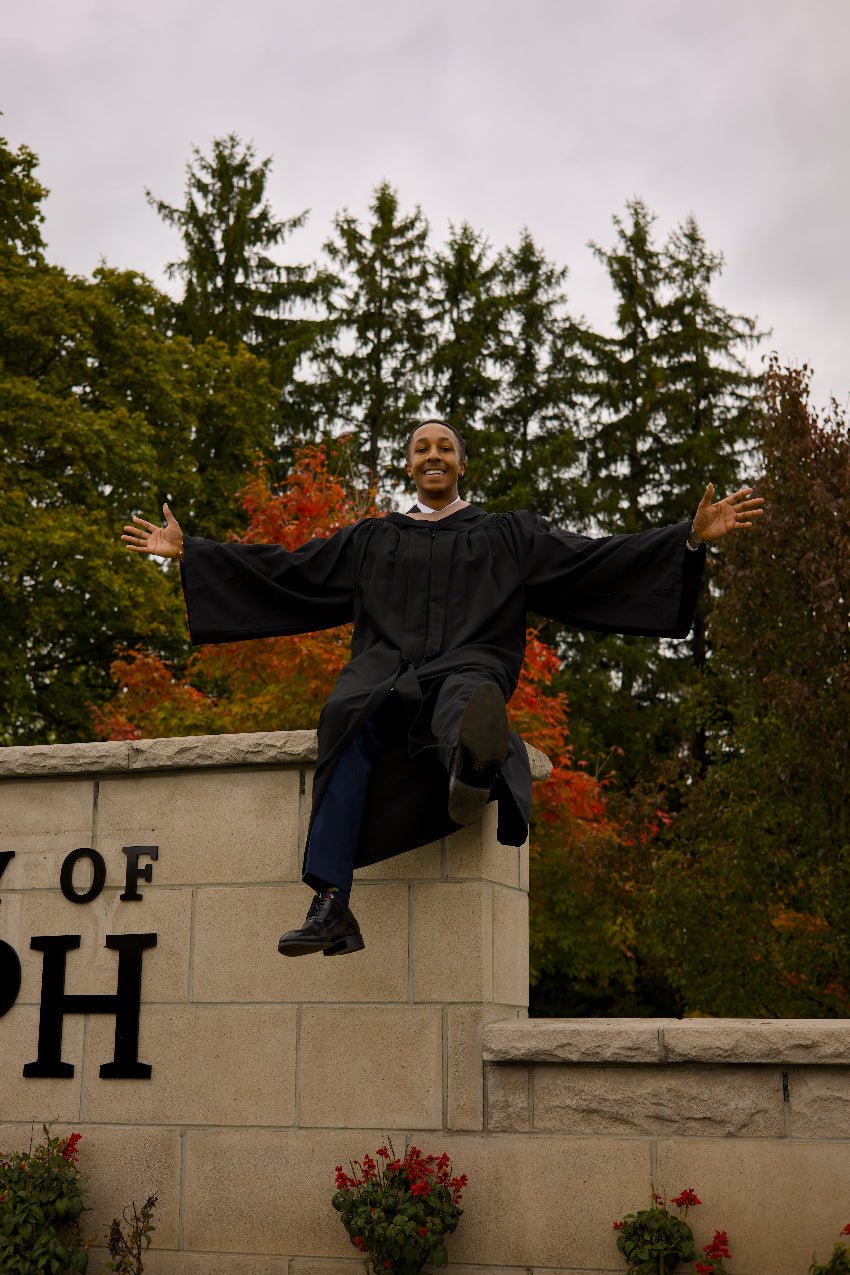 Person in a graduation gown sitting on a stone wall with trees and 'University of ...' sign in the background.