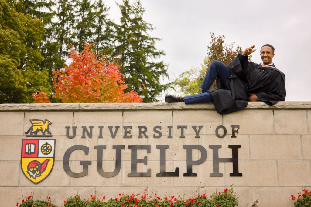 Person in graduation gown sitting on the University of Guelph sign with trees and flowers in the background. The photo is from Guelph Grad Photos. The background is the University of Guelph Campus.