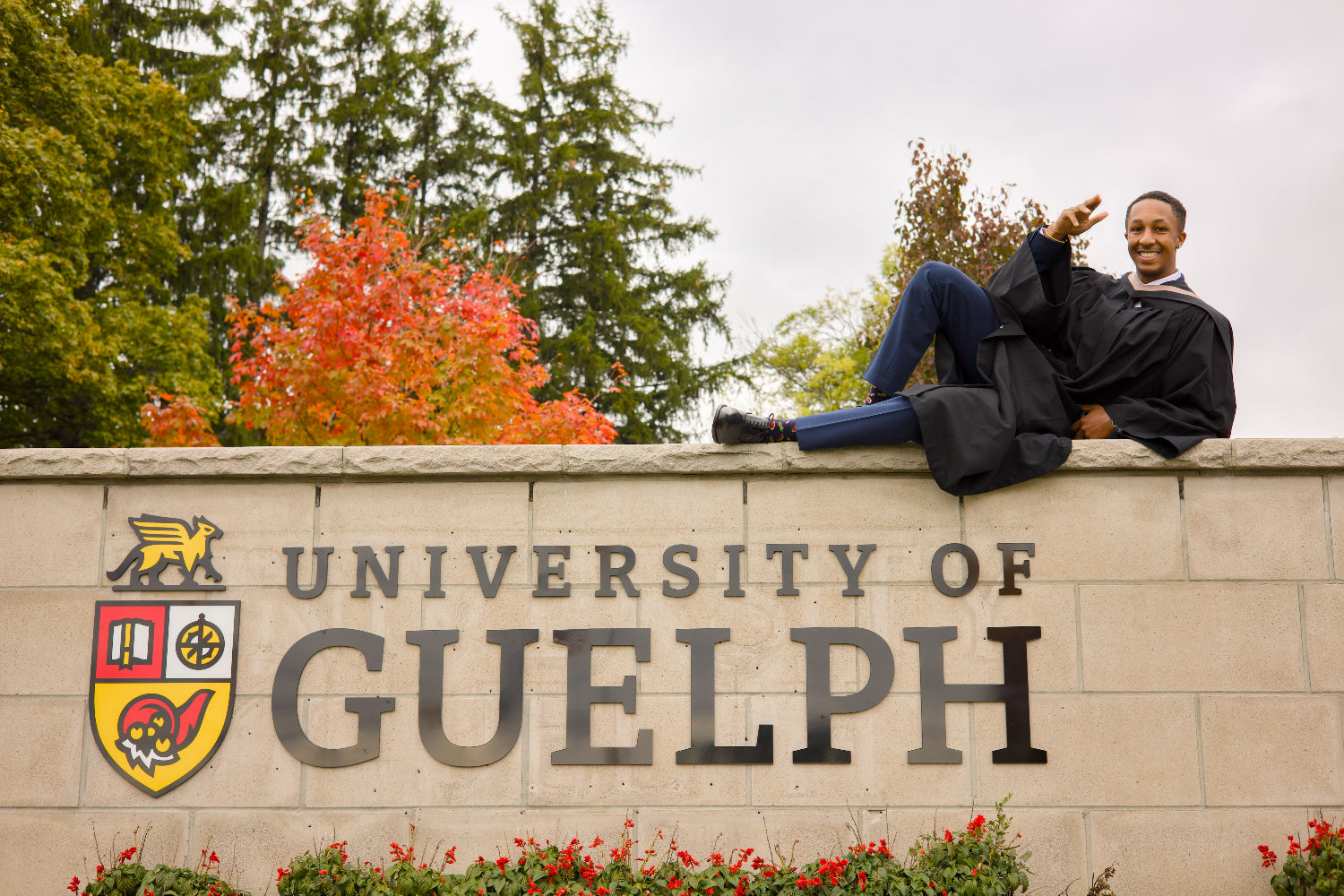 Person in graduation gown sitting on the University of Guelph sign with trees and flowers in the background. The photo is from Guelph Grad Photos. The background is the University of Guelph Campus.