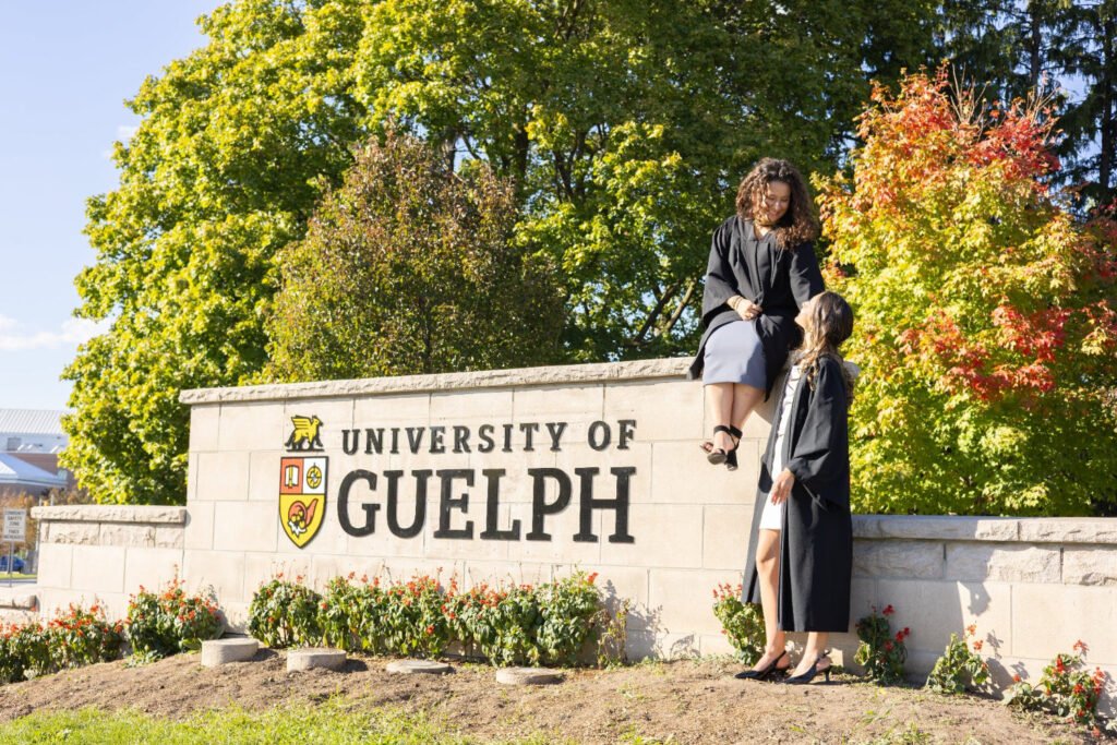 Two people sitting on the University of Guelph sign with trees and flowers in the background. The photo is from Guelph Grad Photos. The background is the University of Guelph Campus.