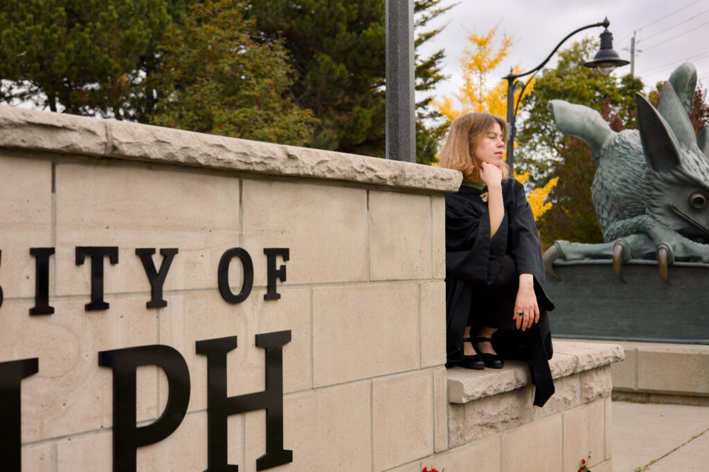 Woman sitting on a stone wall with 'City of Ralph' sign and dragon statue in the background. The photo is from Guelph Grad Photos. The background is the University of Guelph Campus.