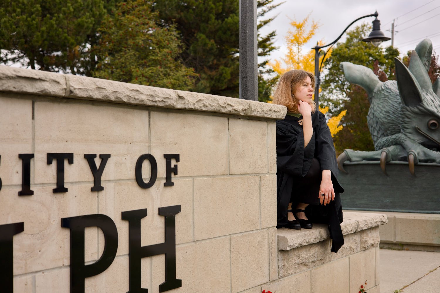 Woman sitting on a stone wall with 'City of Ralph' sign and dragon statue in the background. The photo is from Guelph Grad Photos. The background is the University of Guelph Campus.