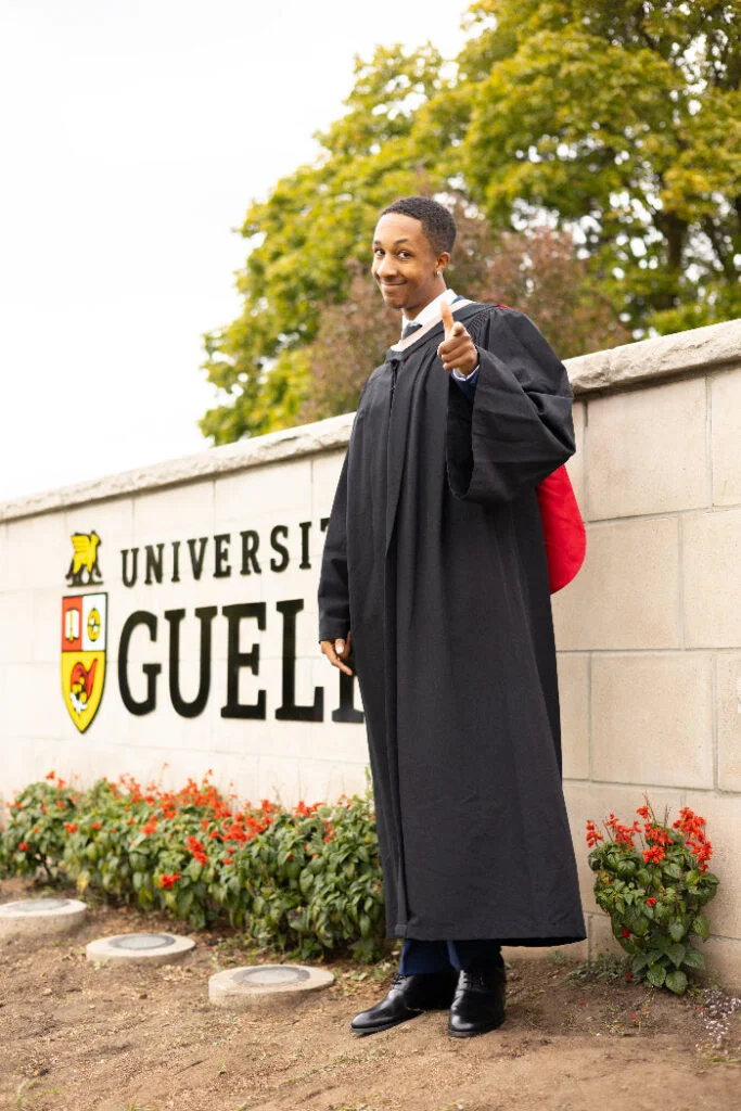 Graduate in cap and gown standing in front of the University of Guelph sign. The photo is from Guelph Grad Photos. The background is the University of Guelph Campus.