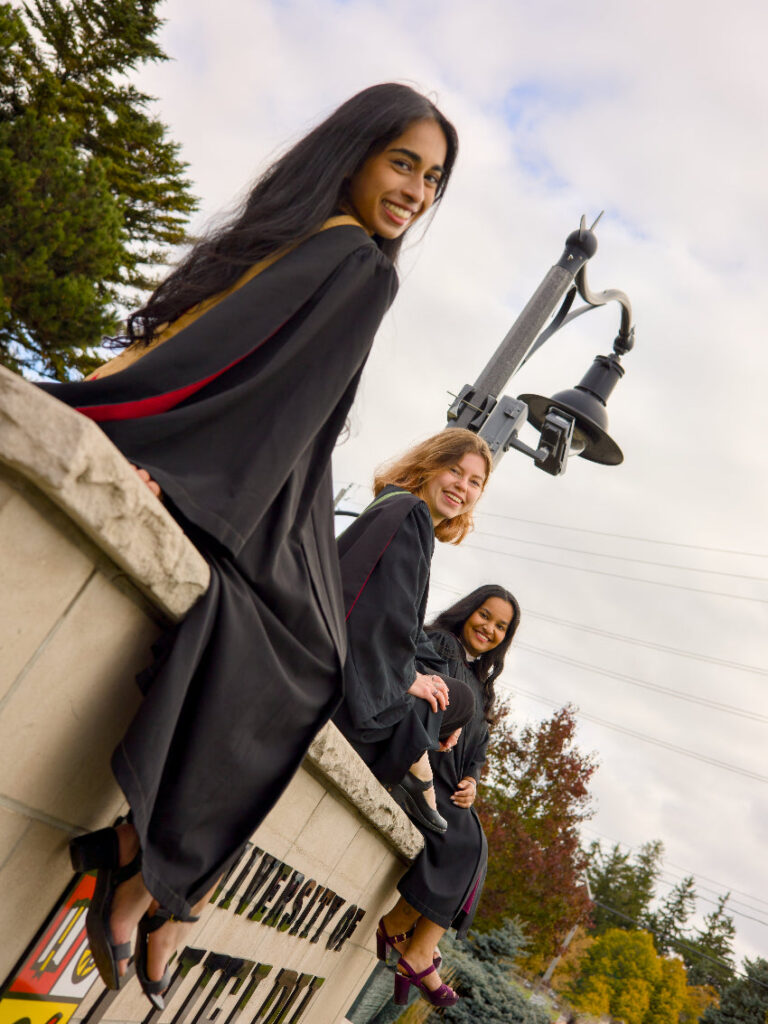 Three women posing on a stone ledge with trees and a light fixture in the background. The photo is from Guelph Grad Photos. The background is the University of Guelph Campus.