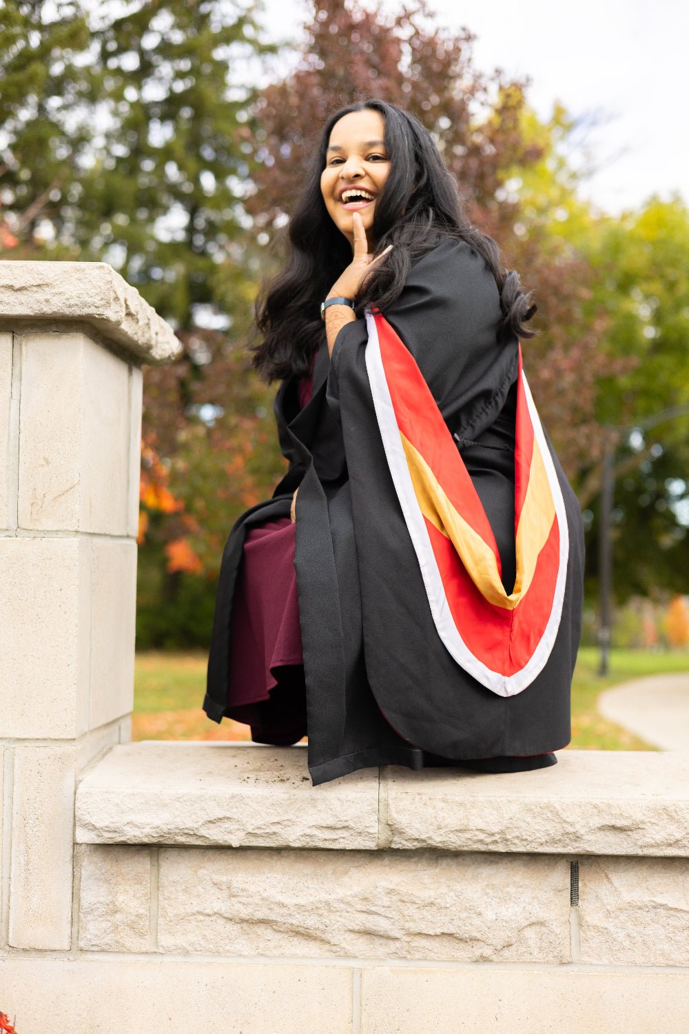 Graduate in cap and gown with a sash sitting on a stone ledge outdoors.