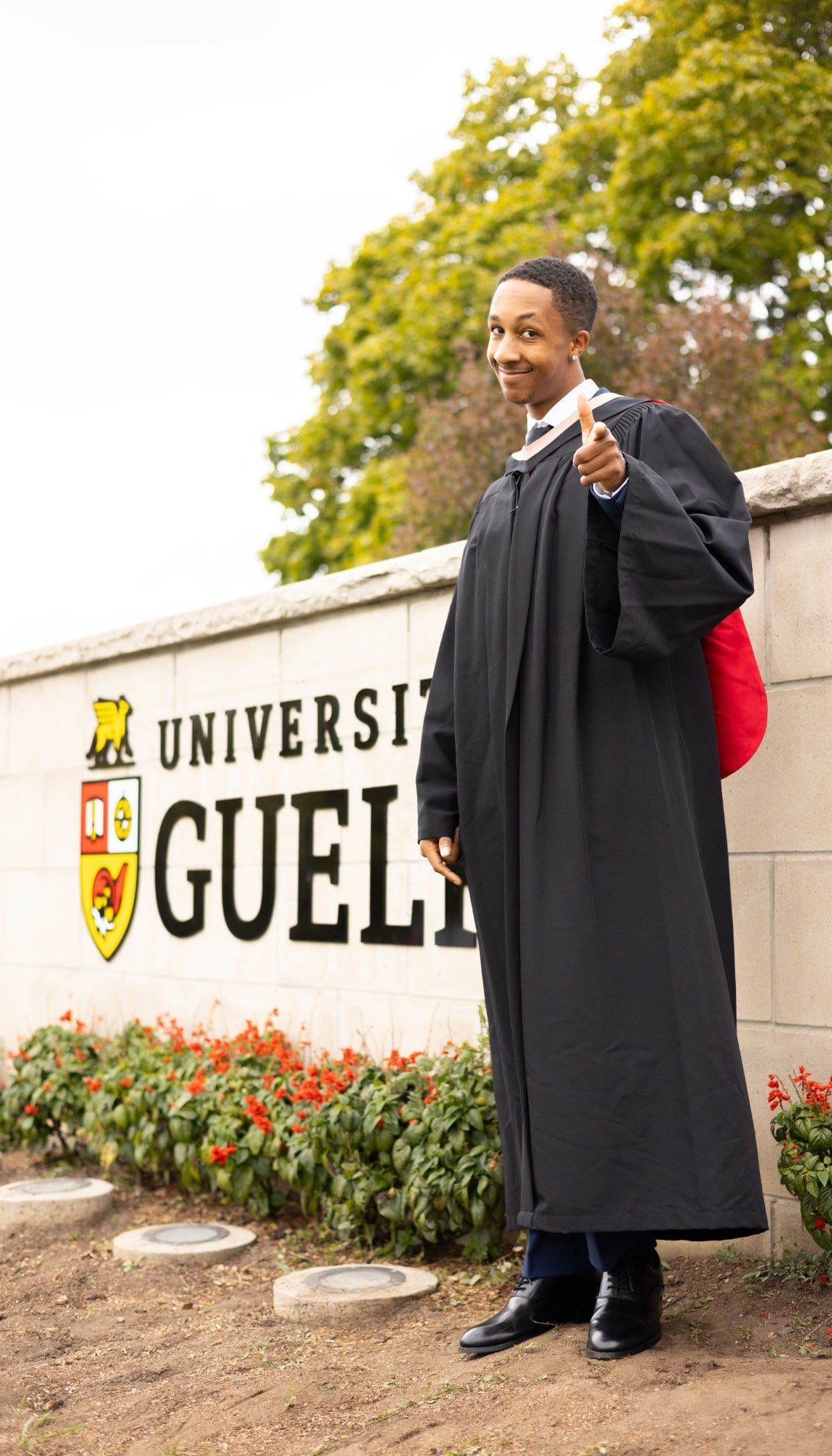 Graduate in cap and gown standing in front of the University of Guelph sign. The photo is from Guelph Grad Photos. The background is the University of Guelph Campus.