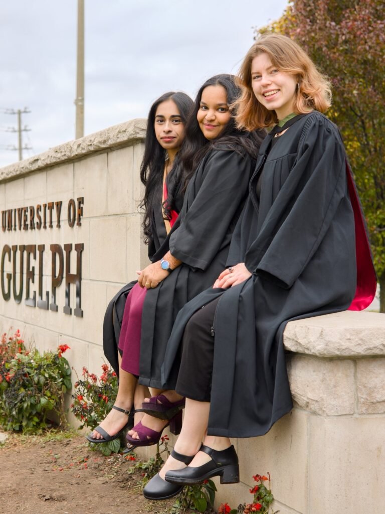 A professional photo of three Guelph grads on the University of Guelph campus in graduation gowns, posing in front of the University of Guelph sign. The photo is from Guelph Grad Photos. The background is the Main Entrance Sign & Stone Gates on the University of Guelph Campus.