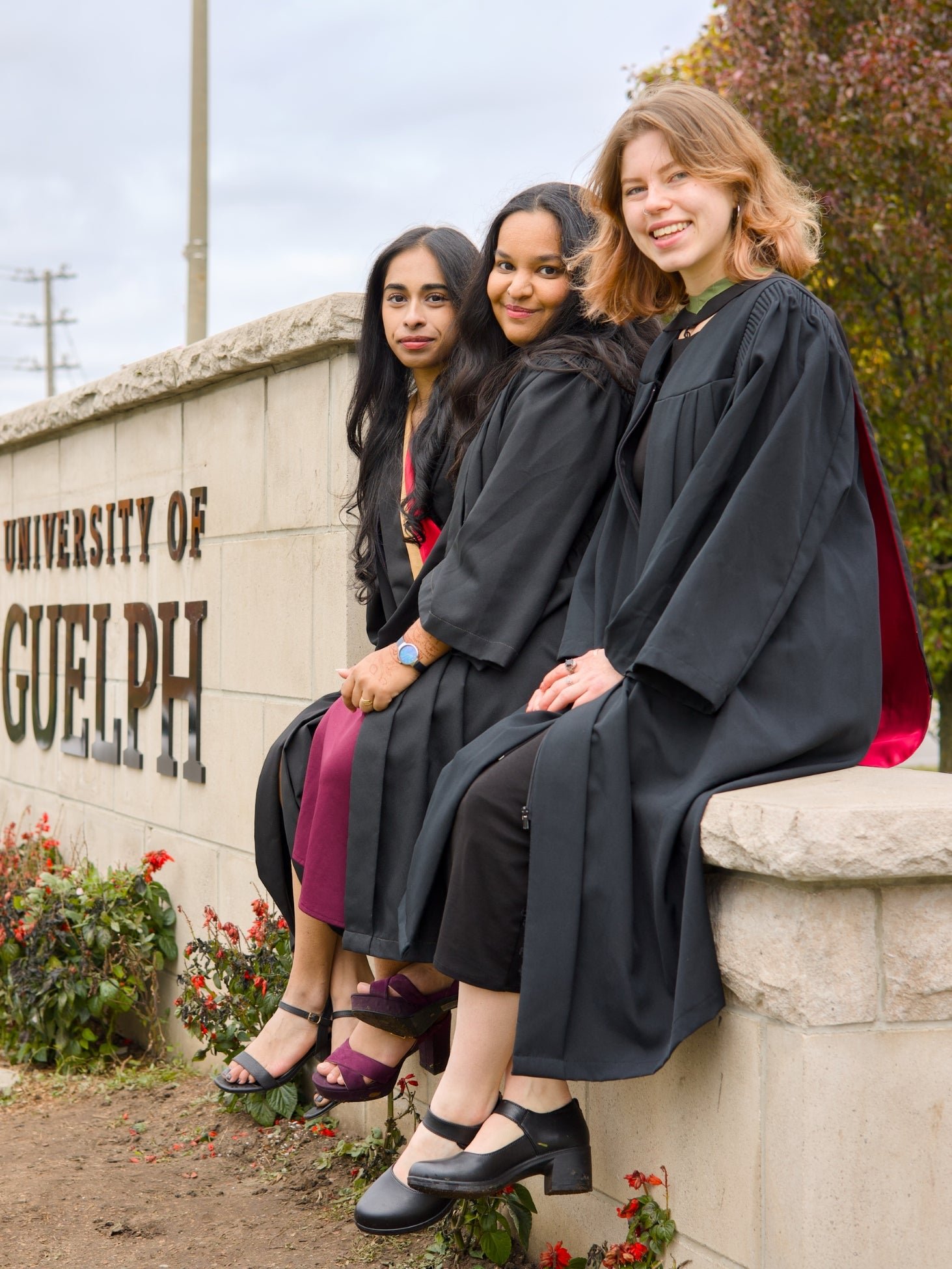 A professional photo of three Guelph grads on the University of Guelph campus in graduation gowns, posing in front of the University of Guelph sign. The photo is from Guelph Grad Photos. The background is the Main Entrance Sign & Stone Gates on the University of Guelph Campus.