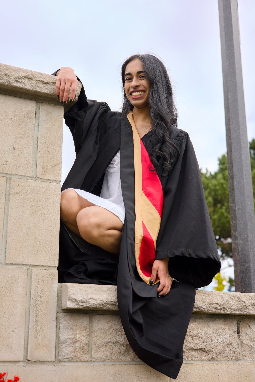 Graduate in cap and gown with a colorful sash sitting on a stone wall. The photo is from Guelph Grad Photos. The background is the University of Guelph Campus.