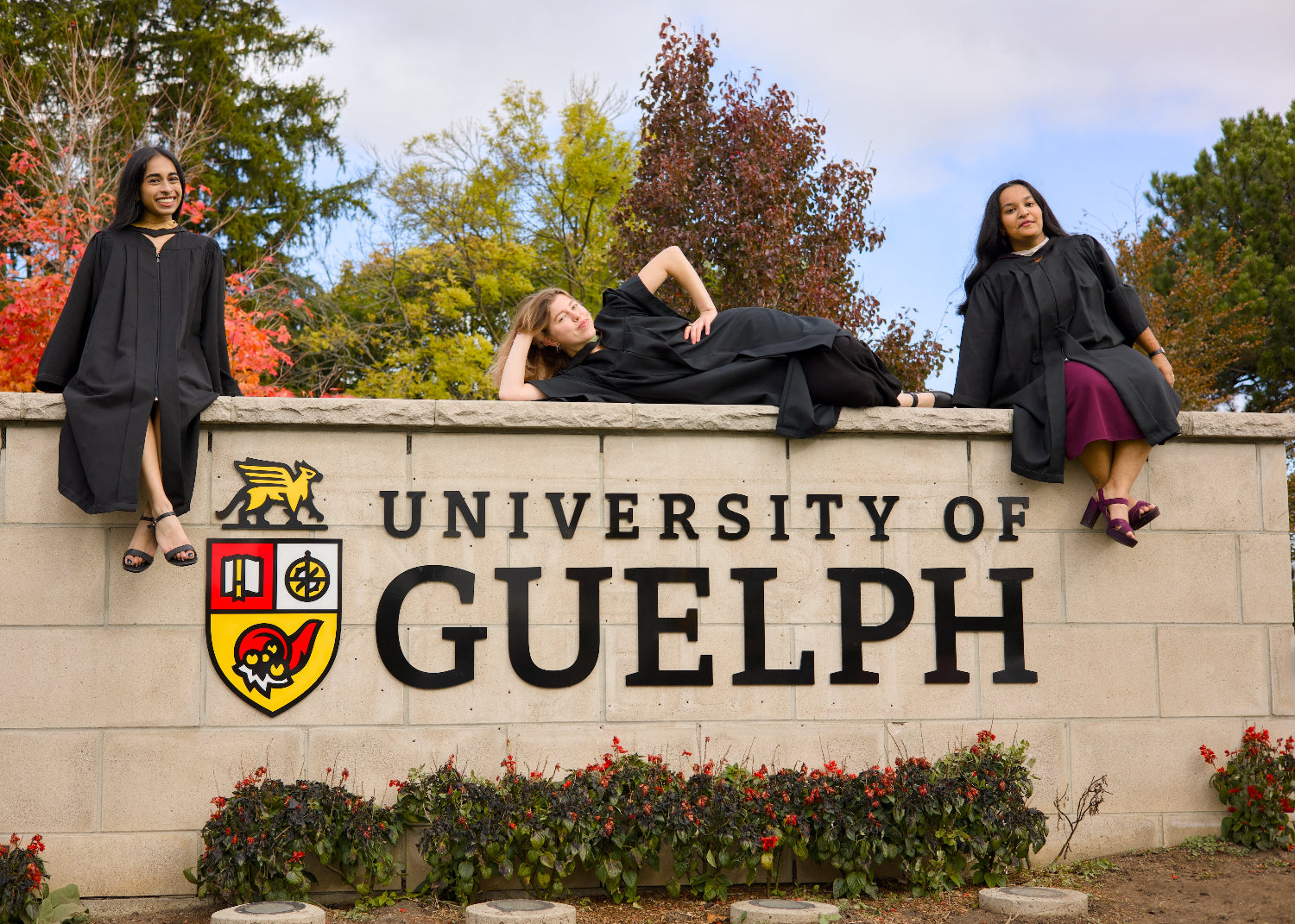 Three individuals in graduation gowns sitting on a wall with the University of Guelph sign in the background. The photo is from Guelph Grad Photos. The background is the University of Guelph Campus.