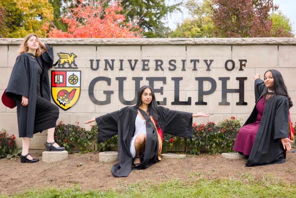 A professional photo of three Guelph grads on the University of Guelph campus in graduation gowns, posing in front of the University of Guelph sign. The photo is from Guelph Grad Photos. The background is the Main Entrance Sign & Stone Gates on the University of Guelph Campus.