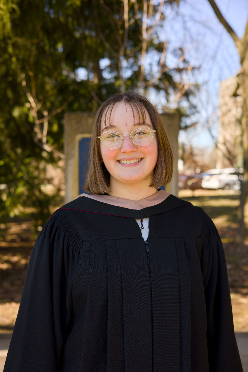 Person in a black graduation gown standing outdoors with trees and a building in the background.