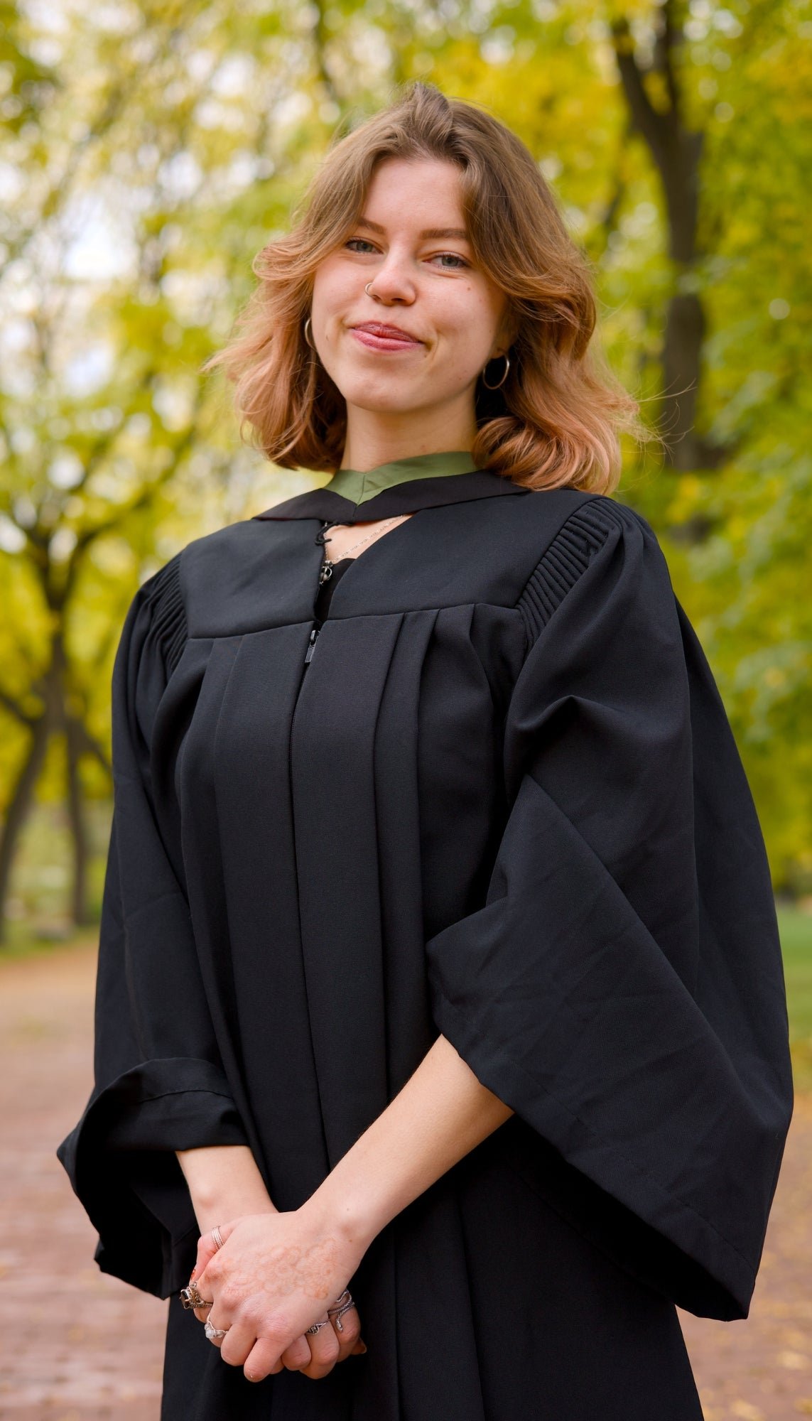 Person in a black graduation gown standing outdoors with trees in the background, The photo is from Guelph Grad Photos. The background is the University of Guelph Campus.