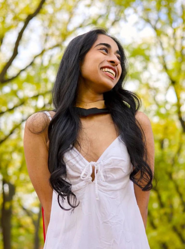 Woman in a white dress standing outdoors with greenery in the background. The photo is from Guelph Grad Photos. The background is the University of Guelph Campus.