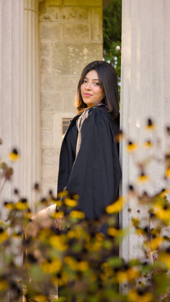 A professional photo of a Guelph grad on the University of Guelph campus. She is in a black coat, standing among yellow flowers with a classical building in the background. The photo is from Guelph Grad Photos. The background is The Portico on the University of Guelph Campus.