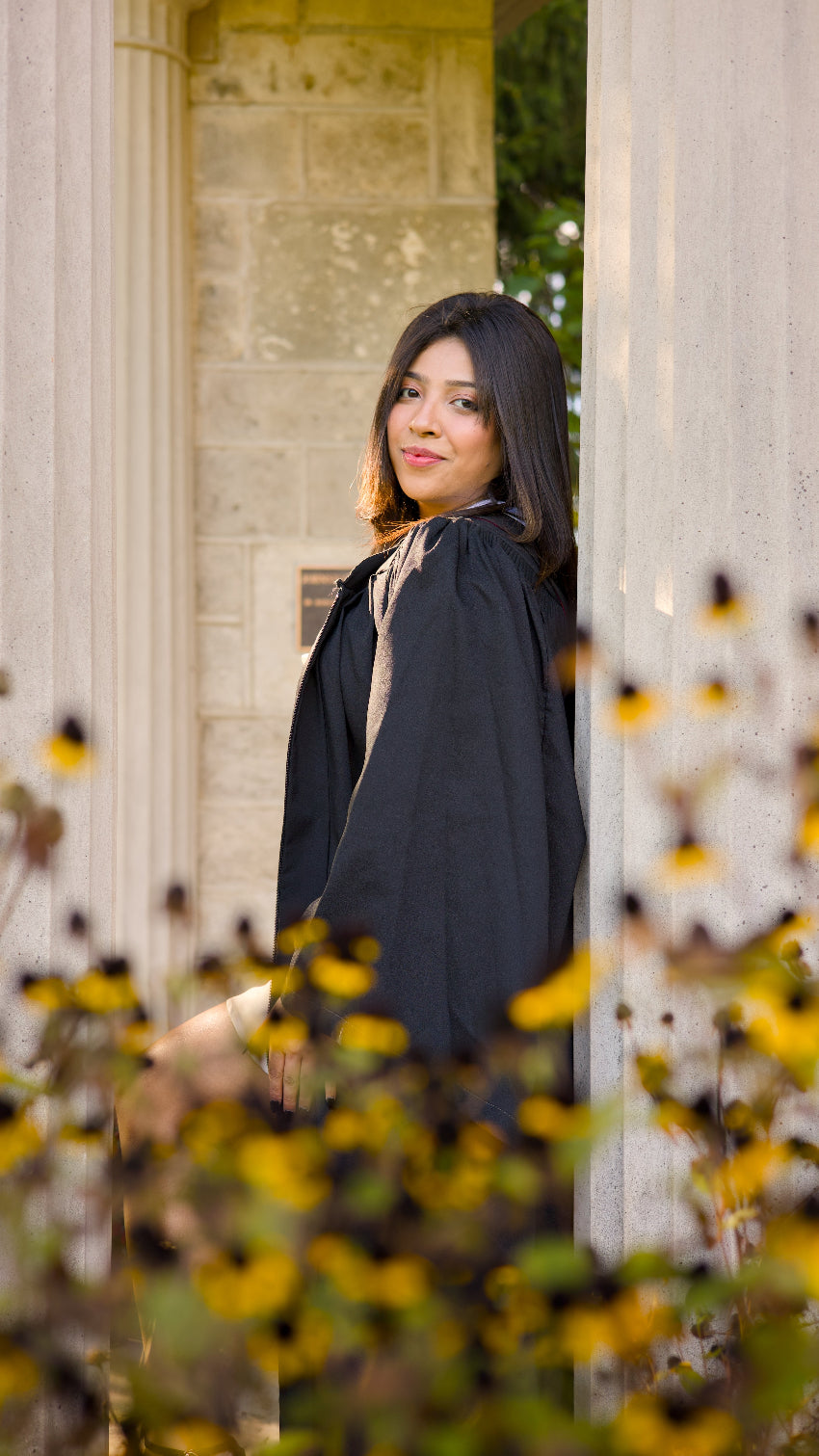 A professional photo of a Guelph grad on the University of Guelph campus. She is in a black coat, standing among yellow flowers with a classical building in the background. The photo is from Guelph Grad Photos. The background is The Portico on the University of Guelph Campus.