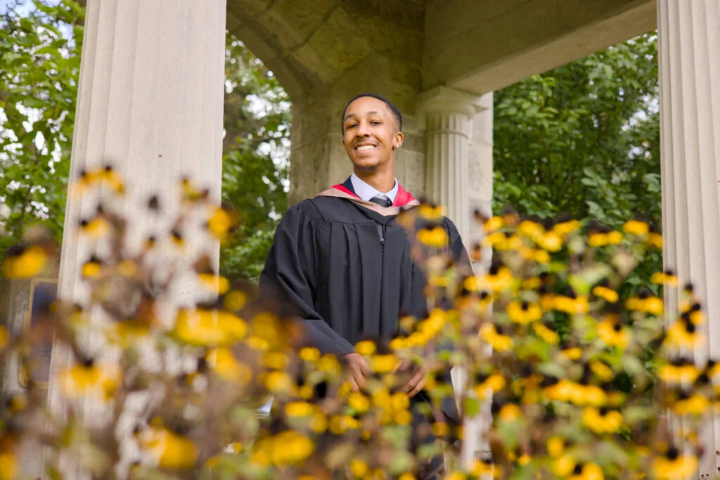 Graduate in cap and gown standing outdoors with columns and yellow flowers in the foreground, The photo is from Guelph Grad Photos. The background is the University of Guelph Campus.