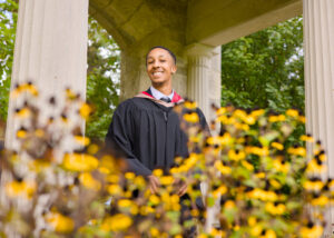 Graduate in cap and gown standing among yellow flowers with classical architecture in the background. The photo is from Guelph Grad Photos. The background is the University of Guelph Campus.