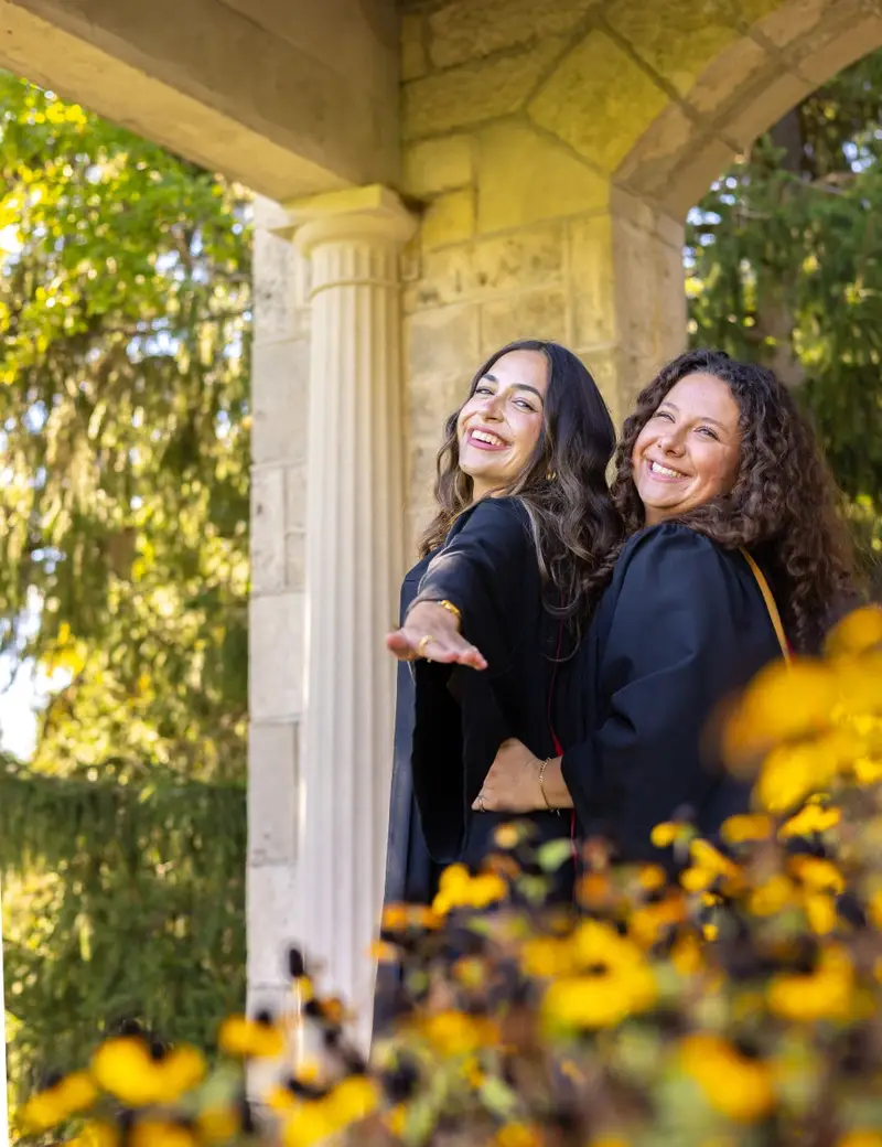 Two women standing under a stone archway with trees and flowers in the background. The photo is from Guelph Grad Photos. The background is the University of Guelph Campus.