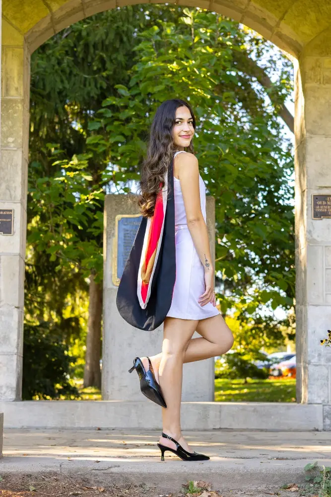 A professional photo of a young woman in a white dress wearing a graduation hood, standing outdoors under a stone archway. The photo is from Guelph Grad Photos. The background is The Portico on the University of Guelph Campus.