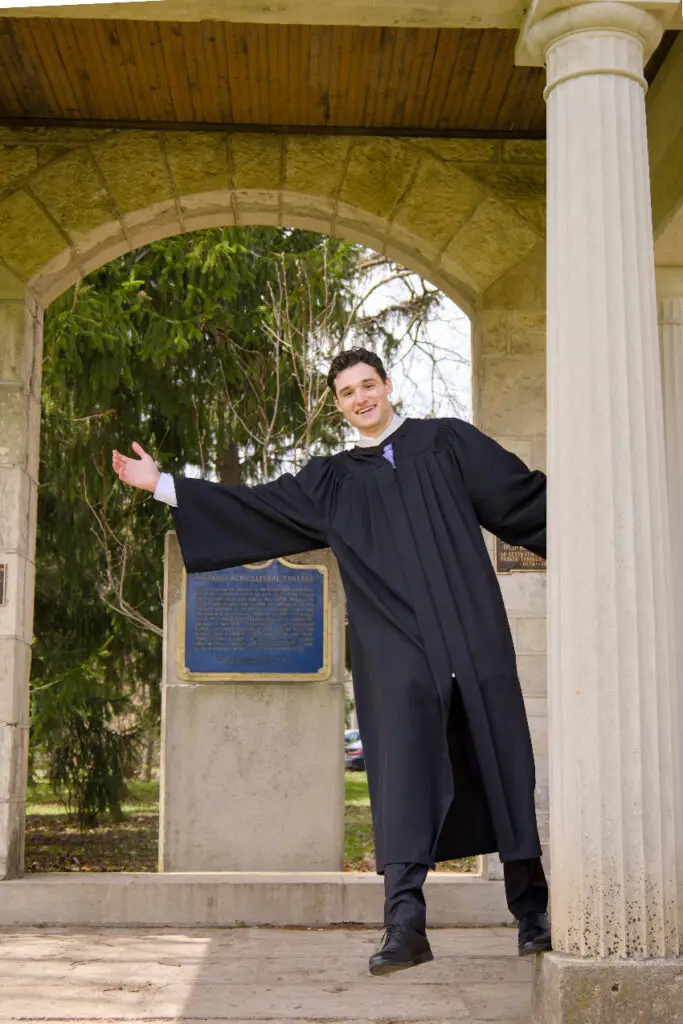 Graduate in a cap and gown standing under an archway with columns. The photo is from Guelph Grad Photos. The background is the University of Guelph Campus.