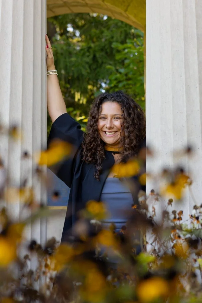 Woman standing behind a white archway with autumn leaves and flowers in the foreground. The photo is from Guelph Grad Photos. The background is the University of Guelph Campus.