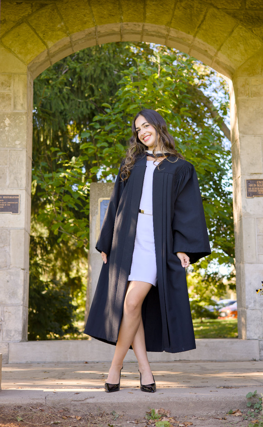Woman in a long dark coat standing in front of an archway with trees in the background. The photo is from Guelph Grad Photos. The background is the University of Guelph Campus.