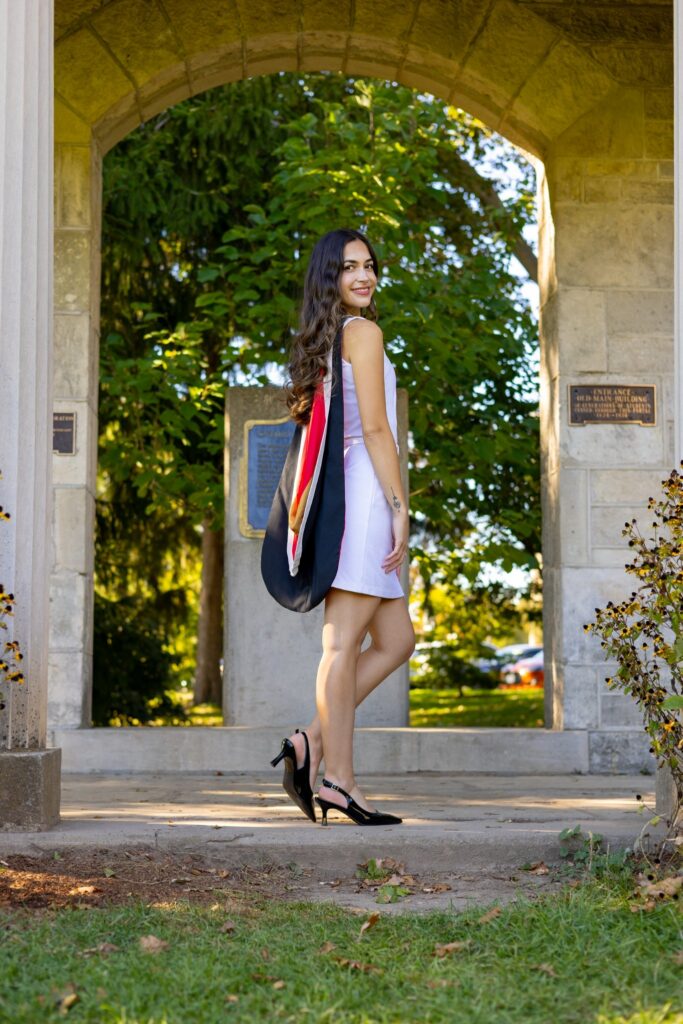 A professional photo of a young woman in a white dress wearing a graduation hood, standing outdoors under a stone archway. The photo is from Guelph Grad Photos. The background is The Portico on the University of Guelph Campus.